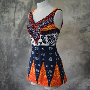 May include: A sleeveless, vintage-style garment with a V-neck, displayed on a mannequin. The top features a vibrant pattern of orange, navy, white, and yellow. The design includes geometric and floral motifs, creating a retro aesthetic.