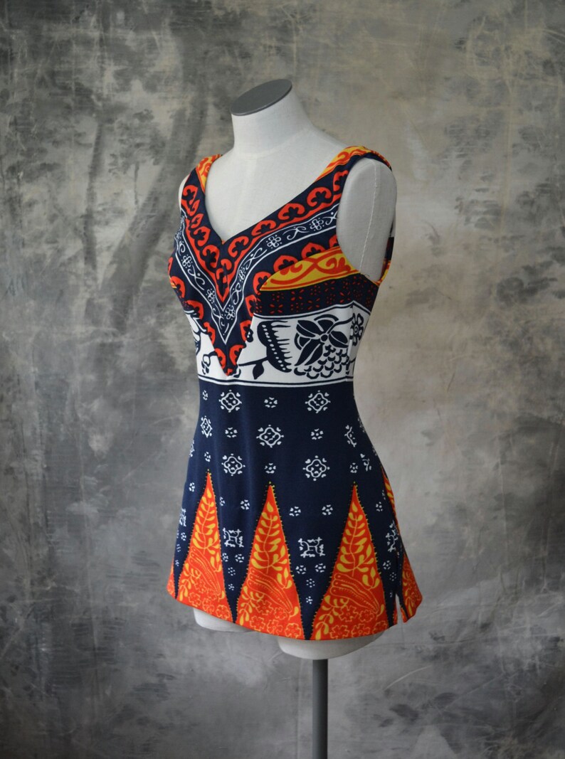 May include: A sleeveless, vintage-style garment with a V-neck, displayed on a mannequin. The top features a vibrant pattern of orange, navy, white, and yellow. The design includes geometric and floral motifs, creating a retro aesthetic.