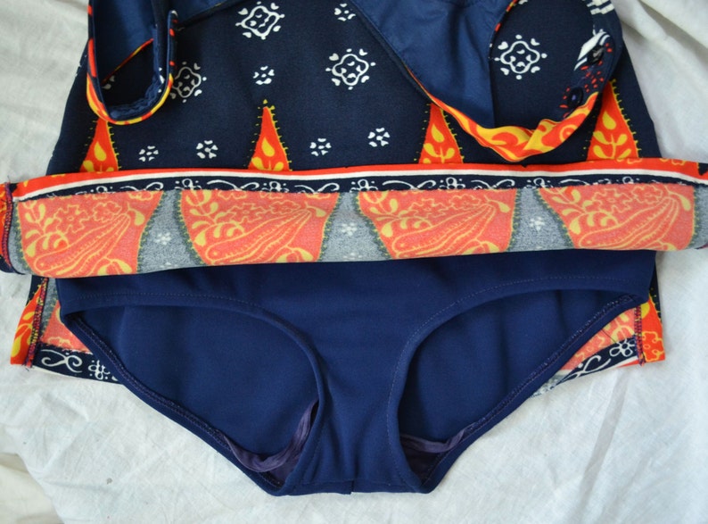 May include: A two-piece swimsuit with a navy blue bottom and a top. The top has a navy blue background and a pattern of white, orange, and yellow shapes. The top has a wrap-around design.