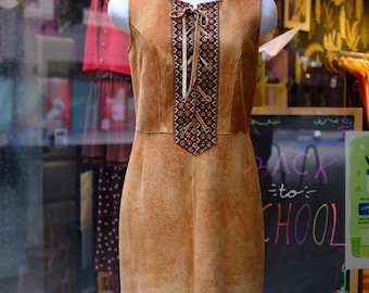 May include: A sleeveless tan suede dress featuring a central panel with a dark blue and orange geometric pattern, a lace-up front closure with tan ties, and a fringe hemline. The dress is displayed on a mannequin in a storefront window.