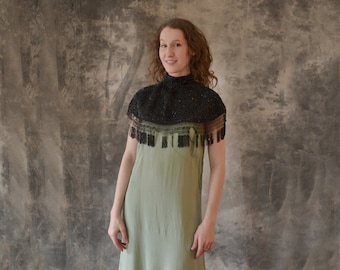 May include: A woman wearing a green dress with a black beaded collar and fringe. The dress has a high neckline and a fitted bodice. The woman is standing in front of a textured background.
