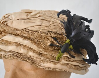 May include: A beige straw hat with a black feather accent. The hat has a layered design with a ruffled edge.