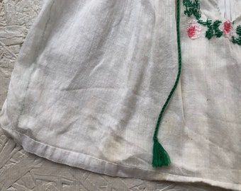 May include: White cotton blouse with a green and pink floral embroidery design. The blouse has a tassel tie at the neck.