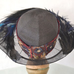 Pode incluir: Um chap&eacute;u de palha preto com aba larga e v&eacute;u de rede preto. O chap&eacute;u &eacute; decorado com uma faixa de tecido vermelho e azul com bordados florais. O chap&eacute;u &eacute; adornado com penas pretas e azuis.