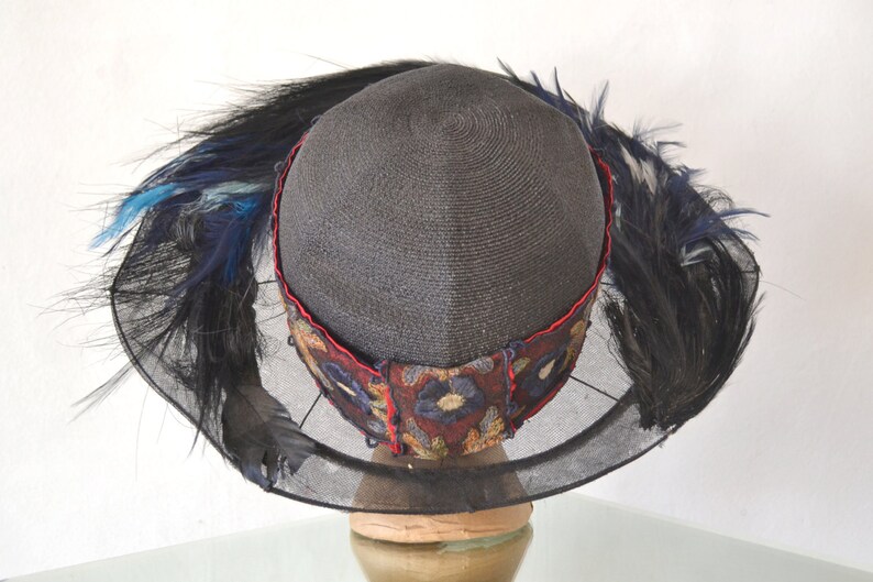 Pode incluir: Um chap&eacute;u de palha preto com aba larga e v&eacute;u de rede preto. O chap&eacute;u &eacute; decorado com uma faixa de tecido vermelho e azul com bordados florais. O chap&eacute;u &eacute; adornado com penas pretas e azuis.