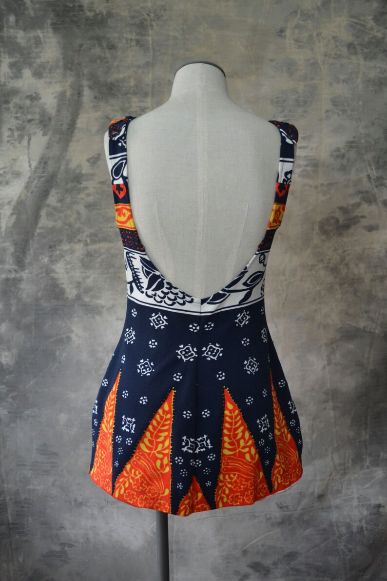 May include: A sleeveless dress with a low back, displayed on a mannequin. The dress has a navy blue base with white, orange, and yellow geometric patterns. The straps and upper back have a contrasting pattern. The hemline features orange and yellow leaf shapes.