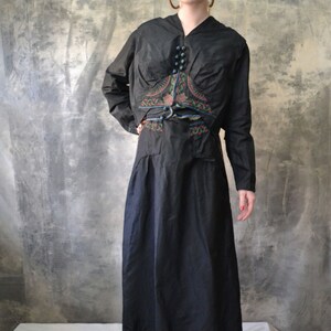 May include: A black long-sleeved dress with a matching jacket. The jacket has a decorative embroidered design with floral and geometric patterns in shades of blue, green, and red. The dress has a fitted bodice and a flowing skirt.
