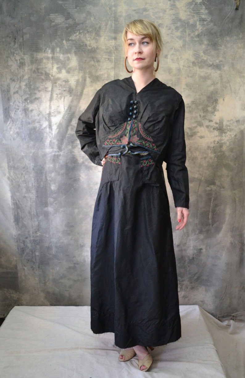 May include: A black long-sleeved dress with a matching jacket. The jacket has a decorative embroidered design with floral and geometric patterns in shades of blue, green, and red. The dress has a fitted bodice and a flowing skirt.
