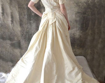 May include: Ivory-colored vintage wedding dress with a short-sleeve bodice and a full skirt. The dress features a low back and a row of buttons down the back. The skirt has a dramatic, flowing shape, creating a classic bridal look.