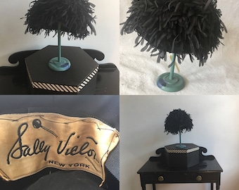 May include: A black feather hat on a display stand with a label that reads "Sally Viéla NEW YORK".