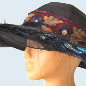 Pode incluir: Um chap&eacute;u preto de aba larga com uma faixa bordada em vermelho, azul e marrom e penas azuis.