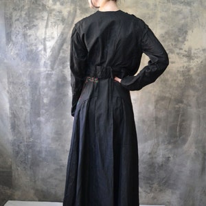 May include: A long black dress with a floral embroidered belt. The dress has long sleeves and a fitted bodice. The dress is photographed from the back.