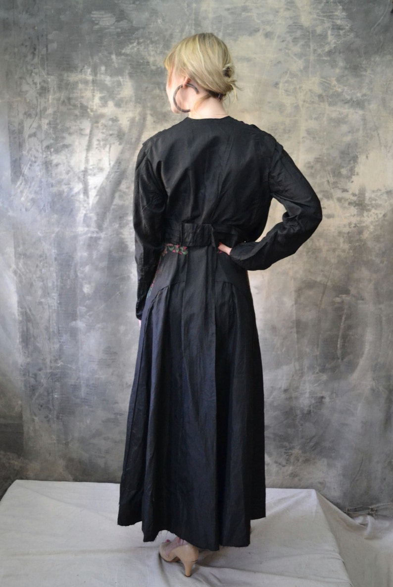 May include: A long black dress with a floral embroidered belt. The dress has long sleeves and a fitted bodice. The dress is photographed from the back.