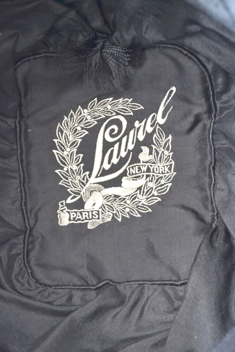 Pode incluir: Um chap&eacute;u de tecido preto com um logotipo Laurel bordado em branco. O logotipo apresenta uma coroa com as palavras "Laurel", "Nova Iorque" e "Paris".