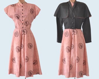 May include: A pink dress with a black floral print and a black jacket with a cape. The dress has a button-up front and a full skirt. The jacket has a button-up front and a cape collar.