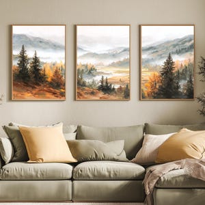 May include: Three framed landscape paintings showing a misty mountain scene with trees in green, orange, and brown hues. The artwork is displayed above a green sofa with yellow and white cushions, creating a warm atmosphere.