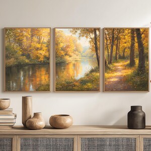 Könnte beinhalten: Triptychon gerahmter Landschaftsbilder, die eine Herbstszene mit einem Fluss und einem Weg durch einen Wald zeigen. Die Gemälde weisen warme Farben auf, darunter Gold, Gelb und Braun. Dekorative Vasen und Bücher stehen auf einem Holzschrank.