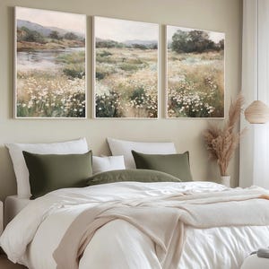 Op de afbeelding: Een slaapkamer met een bed met wit beddengoed, olijfgroene kussens en een beige deken. Boven het bed hangen drie ingelijste landschapsschilderijen van velden en een rivier. De kamer heeft een neutrale kleurenschema.