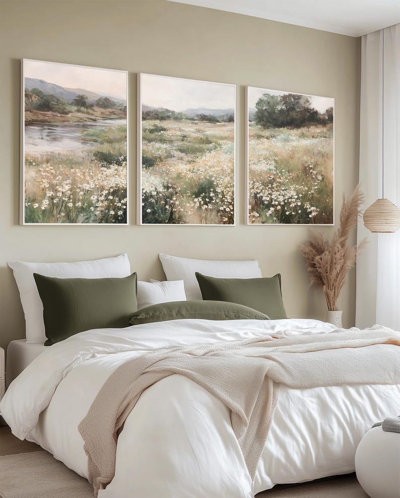 Op de afbeelding: Een slaapkamer met een bed met wit beddengoed, olijfgroene kussens en een beige deken. Boven het bed hangen drie ingelijste landschapsschilderijen van velden en een rivier. De kamer heeft een neutrale kleurenschema.