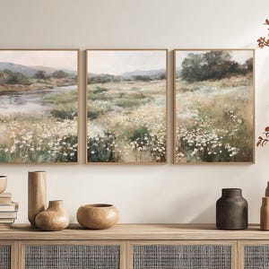 Op de afbeelding: Een drieluik van ingelijste landschapsschilderijen die een serene weidesc&egrave;ne met een rivier en wilde bloemen uitbeelden. Het kunstwerk wordt tentoongesteld boven een houten kast met decoratieve vazen ​​en boeken.