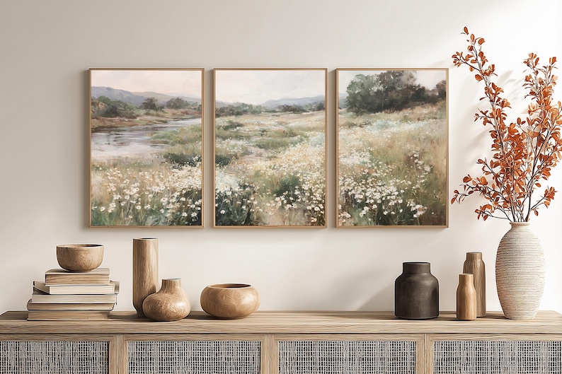 Op de afbeelding: Een drieluik van ingelijste landschapsschilderijen die een serene weidesc&egrave;ne met een rivier en wilde bloemen uitbeelden. Het kunstwerk wordt tentoongesteld boven een houten kast met decoratieve vazen ​​en boeken.
