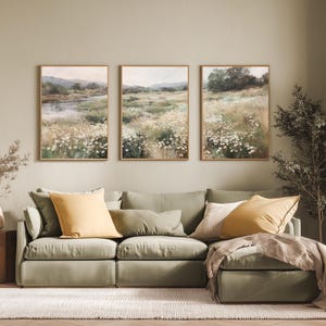 Op de afbeelding: Drie ingelijste landschapsschilderijen met velden vol wilde bloemen en verre heuvels. Het kunstwerk gebruikt een gedempt kleurenpalet van groen, wit en bruin, met houten frames. De schilderijen worden boven een groene bank met gele en cr&egrave;mekleurige kussens getoond.