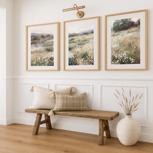 Op de afbeelding: Drie ingelijste landschapsschilderijen met gouden frames hangen boven een houten bank met kussens. De schilderijen tonen velden met wilde bloemen. Een vaas met gedroogde grassen staat op de grond. De bank heeft een lichtgekleurd kussen met kwastjes en een geruit kussen.