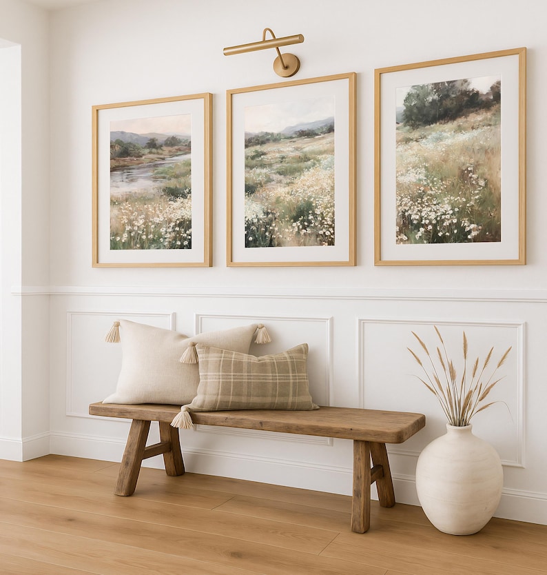 Op de afbeelding: Drie ingelijste landschapsschilderijen met gouden frames hangen boven een houten bank met kussens. De schilderijen tonen velden met wilde bloemen. Een vaas met gedroogde grassen staat op de grond. De bank heeft een lichtgekleurd kussen met kwastjes en een geruit kussen.