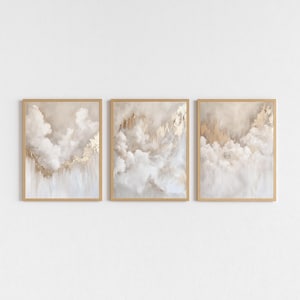 Peut inclure: Ensemble de trois impressions d'art encadrées. Chaque impression présente un design abstrait avec des tons blancs, beiges et dorés, ressemblant à des nuages et des accents métalliques. Les cadres sont de couleur bois clair.