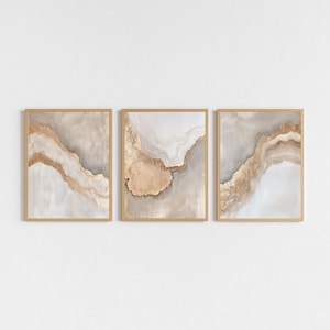 May include: Set of three framed abstract art prints. Each print features a flowing design with shades of beige, gold, and gray, resembling natural stone formations. The artwork is encased in a light brown wooden frame, creating a cohesive display.
