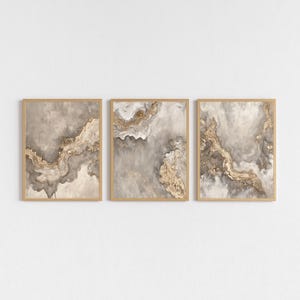 Könnte beinhalten: Drei gerahmte abstrakte Kunstdrucke. Jeder Druck zeigt ein wirbelndes Design in Grau-, Beige- und Goldtönen. Das Kunstwerk ist in hellem Braun gerahmt.
