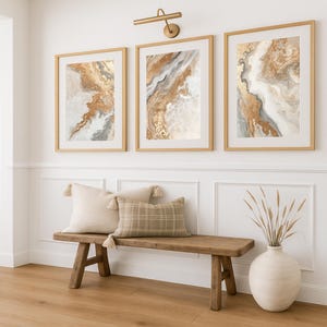 Op de afbeelding: Drie ingelijste abstracte kunstprints met goud-, beige- en grijstinten hangen boven een houten bank met kussens. De kunstwerken tonen vloeiende ontwerpen. Boven de kunst bevindt zich een goudkleurige wandlamp. Een vaas met gedroogde planten staat op de vloer.