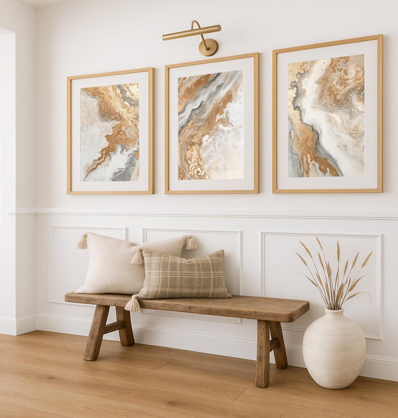 Op de afbeelding: Drie ingelijste abstracte kunstprints met goud-, beige- en grijstinten hangen boven een houten bank met kussens. De kunstwerken tonen vloeiende ontwerpen. Boven de kunst bevindt zich een goudkleurige wandlamp. Een vaas met gedroogde planten staat op de vloer.