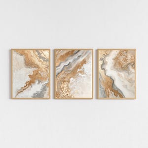 Op de afbeelding: Drie ingelijste abstracte kunstprints. Elke print heeft een vloeiend ontwerp met goud-, wit- en grijstinten. Het kunstwerk is ingelijst in een lichtbruine houten lijst, waardoor een samenhangende set voor wanddecoratie ontstaat.