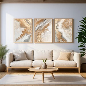 Op de afbeelding: Drie ingelijste abstracte kunstprints met goud-, beige- en grijstinten hangen boven een cr&egrave;mekleurige bank. Een ronde houten salontafel en een plant in pot staan voor de bank. De kamer heeft een modern, minimalistisch ontwerp.