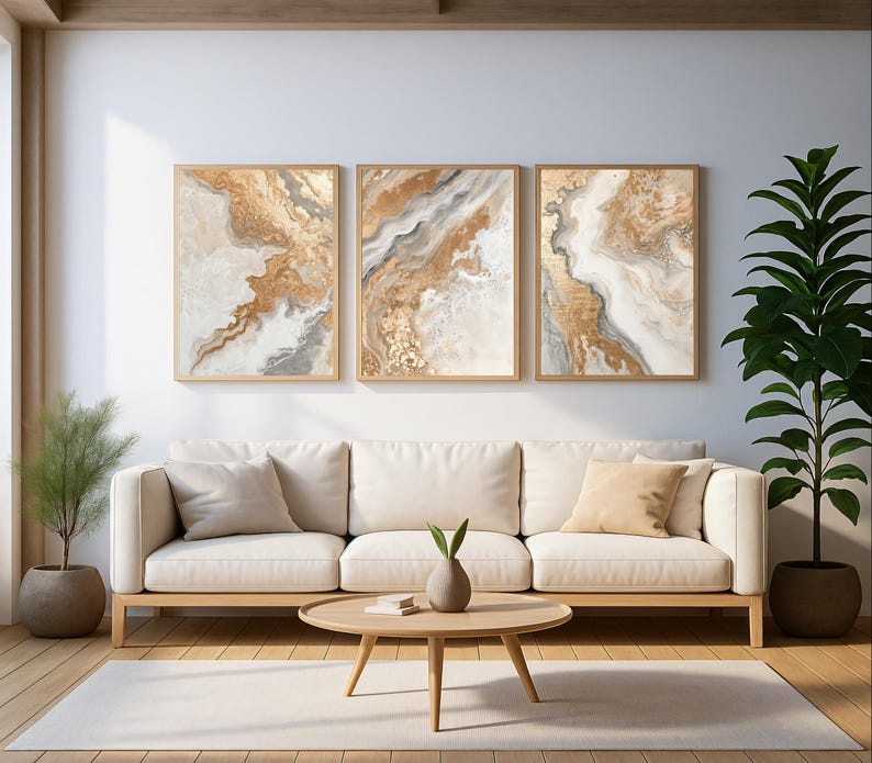Op de afbeelding: Drie ingelijste abstracte kunstprints met goud-, beige- en grijstinten hangen boven een cr&egrave;mekleurige bank. Een ronde houten salontafel en een plant in pot staan voor de bank. De kamer heeft een modern, minimalistisch ontwerp.