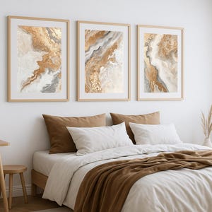 Op de afbeelding: Drie ingelijste abstracte kunstprints in goud, bruin en wit hangen boven een bed met wit beddengoed en bruine kussens en een sprei. Het kunstwerk heeft een vloeiend, gemarmerd ontwerp. De kamer heeft een neutraal kleurenschema.
