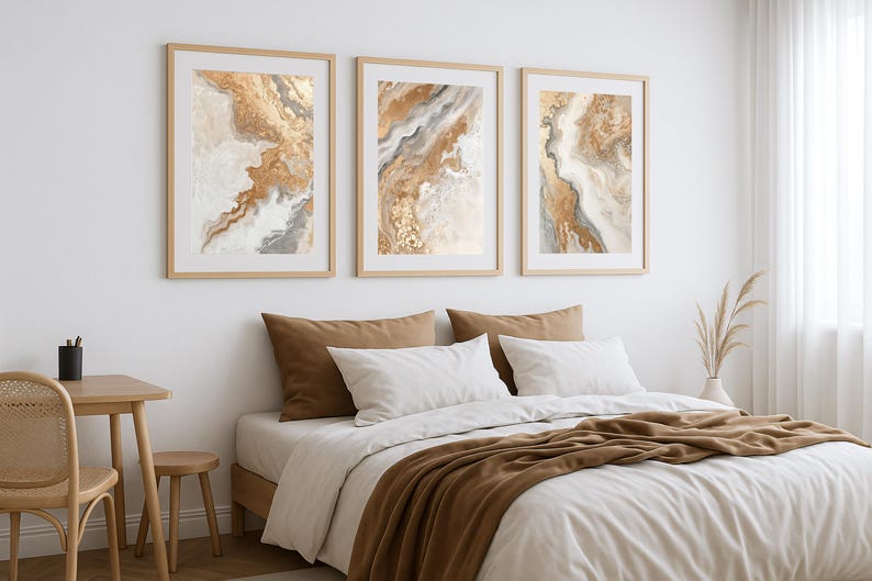 Op de afbeelding: Drie ingelijste abstracte kunstprints in goud, bruin en wit hangen boven een bed met wit beddengoed en bruine kussens en een sprei. Het kunstwerk heeft een vloeiend, gemarmerd ontwerp. De kamer heeft een neutraal kleurenschema.