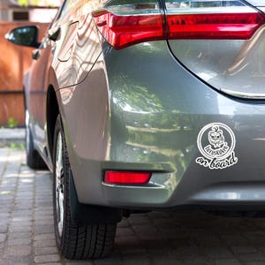 May include: A gray car with a white sticker that says "Luxuskuken on board." The sticker features a cartoon face inside a circle. The car's taillight is red.