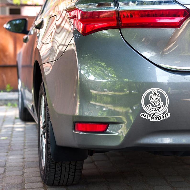 May include: A gray car with a white sticker that says "Luxuskuken on board." The sticker features a cartoon face inside a circle. The car's taillight is red.