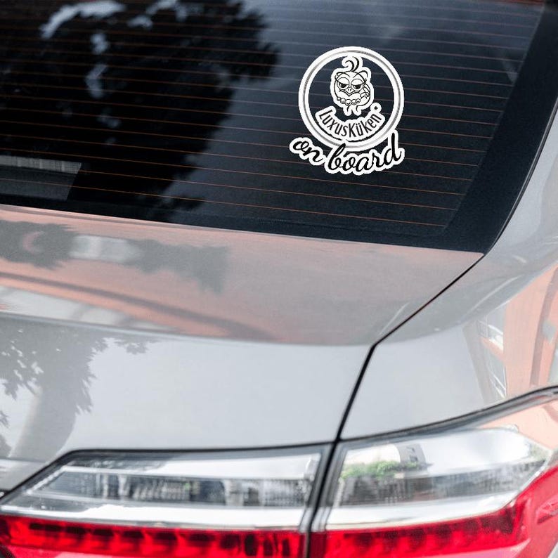 May include: White sticker with the text "LuxusKuken on board" in a stylized font. The sticker features a cartoon character inside a circular frame. The sticker is affixed to the rear window of a silver car.