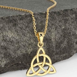 May include: A gold-colored pendant necklace featuring a Celtic knot design. The pendant is a three-part knot, suspended from a delicate chain. The necklace is displayed against a textured, gray background.