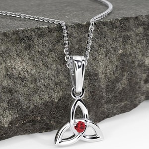 May include: A silver pendant necklace with a Celtic trinity knot design. The pendant features three interlocking arcs and a small, round, red gemstone at the centre. The delicate silver chain rests on a grey stone surface.