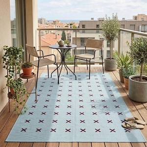 Puede incluir: Una alfombra rectangular de color azul claro con un patrón repetitivo de pequeñas marcas en forma de x de color rojo oscuro, colocada sobre un suelo de balcón de madera bajo una mesa de bistró redonda de metal, dos sillas tejidas y varias plantas.
