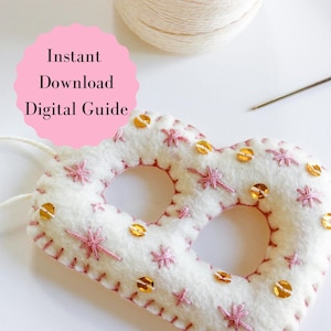 May include: A handmade, white felt pretzel ornament with pink embroidered starbursts and gold sequins. The ornament has a stitched border and is shown with a spool of thread, a needle, and text that reads "Instant Download Digital Guide."