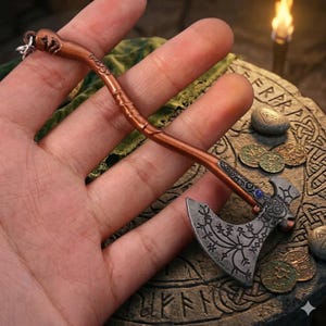 Könnte beinhalten: Miniaturanhänger in Form einer bronzefarbenen Axt mit aufwendigen Details. Der Axtkopf hat eine silberne Oberfläche mit eingravierten Mustern und einem blauen Edelstein. Der Griff ist kupferfarben. Ein kleiner Schlüsselring ist befestigt.