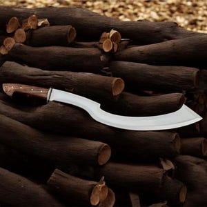 May include: A large, curved blade knife with a wooden handle and a silver-colored blade. The knife is resting on a pile of dark brown logs. The handle is a light brown color.