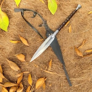 May include: A spear with a silver blade and a brown handle with silver accents, resting in a black leather sheath. The spear is lying on a bed of dry grass and fallen leaves.