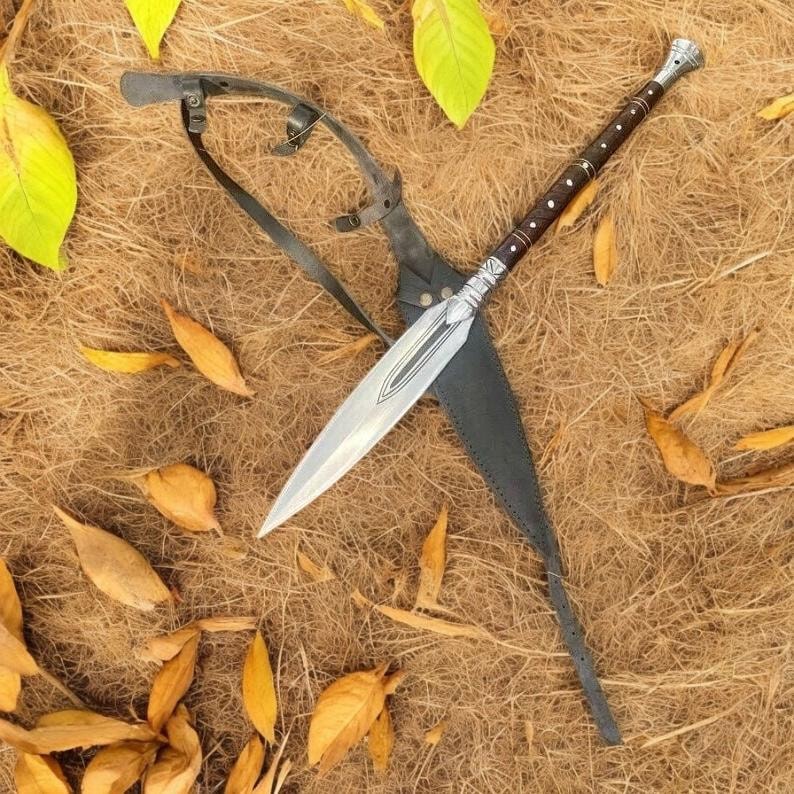 May include: A spear with a silver blade and a brown handle with silver accents, resting in a black leather sheath. The spear is lying on a bed of dry grass and fallen leaves.