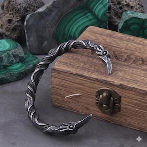 Puede incluir: Una pulsera de puño de metal plateado con un diseño de cuerda trenzada. Los extremos de la pulsera tienen forma de cabezas de pájaro estilizadas con un pequeño ojo de piedra oscura. La pulsera se exhibe en una caja de madera.